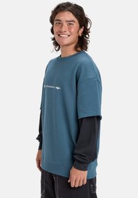 Sudadera de gran tamaño en color verde azulado con mangas cortas, que presenta un estampado de logotipo blanco, usada sobre una camiseta negra de mangas largas. Textura de tela suave.