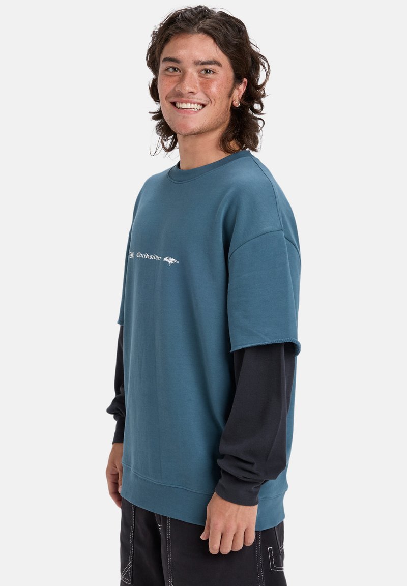 Sudadera de gran tamaño en color verde azulado con mangas cortas, que presenta un estampado de logotipo blanco, usada sobre una camiseta negra de mangas largas. Textura de tela suave.
