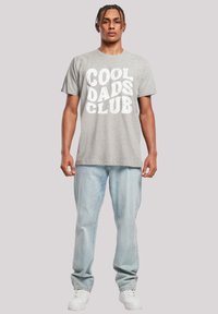 Grijze katoenen T-shirt met opvallende witte tekst "COOL DADS CLUB." Gecombineerd met lichtblauwe denim jeans en witte sneakers.