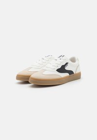 Baskets blanches en cuir avec des accents noirs, zone de bout texturée beige et semelle en caoutchouc gum. Comprend un système de laçage et un col rembourré.
