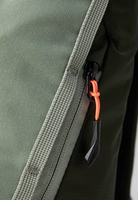 Olivengrønn lerretsryggsekk med kontrasterende glidelås med svart hardware og en oransje trekketapp; teksturert overflate med strukturerte sømmer.