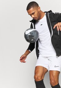 Zwarte zip-up jas met witte accenten, wit t-shirt, witte shorts en zwarte sokken. Een zwart-witte voetbal wordt juggled.