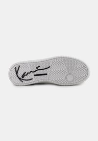 Talpa de pantof athletic alb cu un model de cauciuc texturat, având un accent negru cu semnătura și un logo circular pe toc.