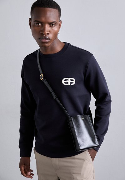 Tengerészkék pulóver logó részlettel; fekete bőr keresztpántos táska sima felülettel; bézs nadrág; a modell előre néz.