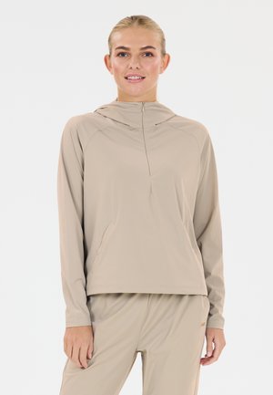 Frau mit blondem Haar, die einen beigefarbenen langärmeligen Kapuzenpullover mit Reißverschluss und passende Hosen trägt, steht vor einem weißen Hintergrund.
