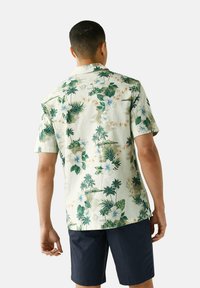 Koszula z krótkim rękawem w jasnym zielonym kolorze, zdobiona kwiatowym wzorem z różnymi tropikalnymi roślinami i kwiatami, wykonana z oddychającego materiału.