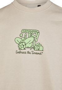 Beige t-shirt featuring a green graphic design of a figure interacting with a soccer ball, text reads "CLEPTOMANIC" and "Embrace the Timeout."
