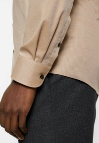 Beige long-sleeve shirt with a textured herringbone pattern, featuring buttoned cuffs and contrasting black buttons; paired with dark textured trousers.