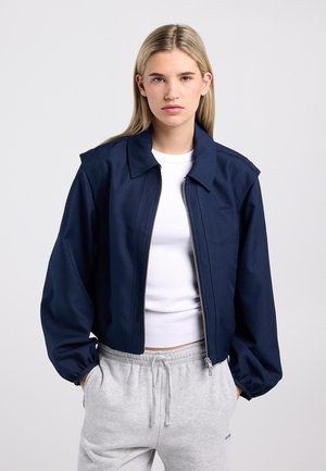 Veste courte bleu marine avec fermeture à glissière, large col et manches bouffantes. Associée à une chemise blanche et un pantalon de survêtement gris.