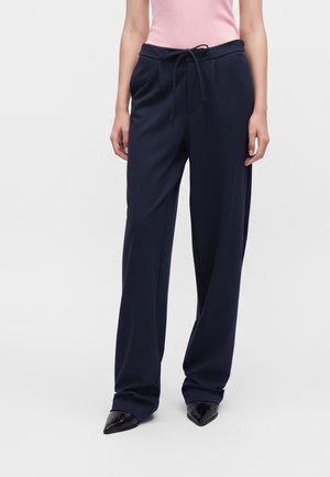 Femme portant un pantalon large bleu marine foncé à taille élastiquée avec cordon et des chaussures noires à bouts pointus, associé à un haut sans manches rose clair.