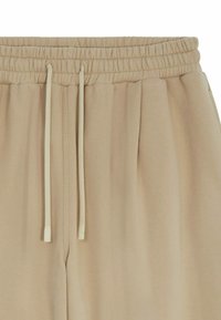 Un pantalon de survêtement beige avec une taille élastique et un cordon de serrage. Le tissu est lisse avec des plis subtils, offrant une coupe décontractée.