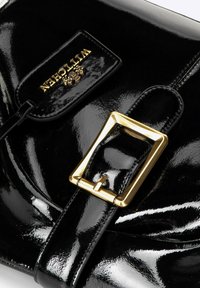 Czarna torebka z lakierowanej skóry z wyraźną złotą kwadratową klamrą i metką z logo. Gładka, błyszcząca tekstura o zorganizowanej konstrukcji.