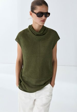 Vrouw draagt een donkergroene mouwloze gebreide top en witte broek met handen in de zakken, geaccentueerd met een donkere zonnebril.
