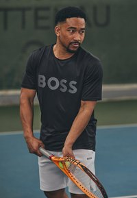 T-shirt de sport noir à manches courtes avec le logo "BOSS", accompagné de shorts blancs. Le joueur tient une raquette de tennis orange avec une balle.
