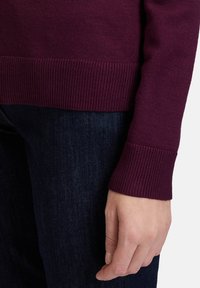 Maglione a maglia color bordeaux con polsini e orlo a coste, caratterizzato da una texture morbida. Indossato con jeans in denim blu scuro.