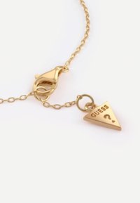 Collana d'oro con chiusura a moschettone, dotata di un ciondolo triangolare inciso con "GUESS" e un punto interrogativo. Finitura in metallo liscia e lucida.