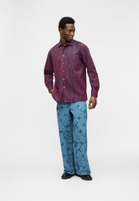 Άντρας που φορά κόκκινο και μπλε πουκάμισο με κάθετες ρίγες, φαρδιά τζιν με μπλε σχέδια και μαύρα παπούτσια, στέκεται έχοντας το ένα του χέρι στην τσέπη.