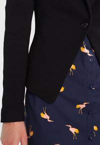 Musta teksturoitu bleiseri, jossa on yksittäinen nappi, puettu tummansinisen mekon päälle, jossa vaaleanpunainen kurkiprintti. Näkyvissä ovat myös ompelusaumat ja napit.