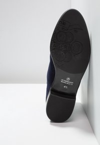 Semelle d'une chaussure habillée noire avec des motifs en relief et une marque Peter Kaiser, taille 4½, appuyée contre un mur blanc d'angle.