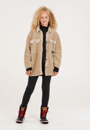 Dijete s kovrčavom plavom kosom koje nosi bež flis jaknu, crne hlače i crne zimske čizme s crvenim vezicama, stoji na jednobojnoj pozadini.