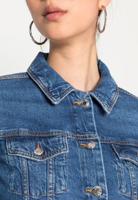 Jeansjacka med blå nyans, med krage, silverfärgade tryckknappar och bröstfickor. Synlig sömnad och texturdetaljer.