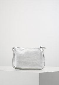 Kleine silberne Metallic-Crossbody-Tasche mit kurzem Riemen, auf einer weißen Oberfläche vor einem schlichten hellgrauen Hintergrund.