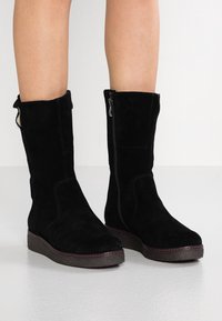 Black suede knee-high boots with a textured surface, rounded toe, and side zipper. Features a thick, contrasting rubber sole.