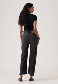 Kvinde med langt krøllet hår iført en sort t-shirt, højtaljede, falmede sorte jeans og sorte slip-on sko, stående med ryggen til.