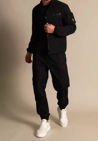 Chaqueta bomber negra con bolsillo con cremallera, combinada con pantalones cargo negros y zapatillas blancas. Textura rugosa en todo el conjunto.