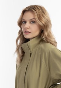 Jonge vrouw met golvend blond haar, gekleed in een hoogsluitende olijfgroene jas, die iets naar de zijkant kijkt tegen een effen witte achtergrond.