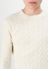 Pullover sweater in cream color, featuring a ribbed crew neckline and textured wave patterns throughout the knit. Soft wool blend fabric.