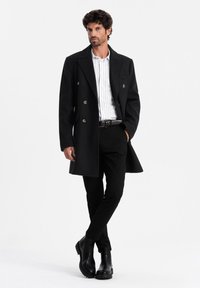 Abrigo negro de corte doble con botones metálicos, ajuste entallado, combinado con una camisa blanca rayada y pantalones negros, usado con botas negras hasta el tobillo.