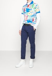 Polo tričko s krátkým rukávem, vícebarevné, s květinovým vzorem, nošené přes bílé tričko s dlouhým rukávem. Námornické kalhoty a bílé tenisky dokončují outfit.