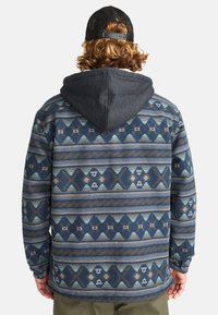 Sweater with a grey hood, fabric shows a geometric blue, brown, and white pattern, featuring distinct shapes and a textured finish.
