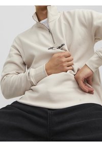 Sudadera de felpa beige con cremallera de cuarto y cuello alzado, que presenta un logo negro, ajuste relajado y puños acanalados, combinada con pantalones negros.