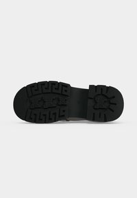 Semelle de chaussure noire avec des motifs de bande de roulement géométriques profonds et des sections antidérapantes texturées sur un fond gris uni.