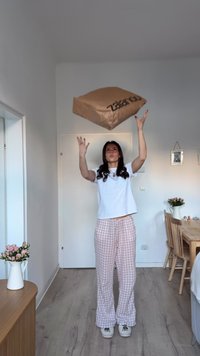 Καφέ χαρτονένιο πακέτο με την επιγραφή «Zalando», κρατημένο πάνω από ένα άτομο που φοράει λευκό μπλουζάκι και ροζ καρό παντελόνι, σε ένα φωτεινό δωμάτιο.