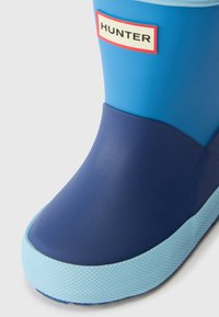 Bota de borracha azul com um acabamento liso, seção inferior em azul mais escuro, sola azul clara e um rótulo de marca na lateral.