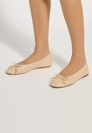 Benedenbenen met beige lakleren ballerina's met kleine strikjes, afgebeeld tegen een effen lichte achtergrond.