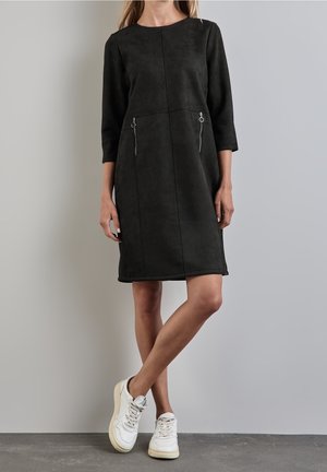 Vrouw draagt een zwarte jurk tot op de knie met driekwart mouwen en ritssluitingdetails, gecombineerd met witte sneakers, staand tegen een grijze muur.