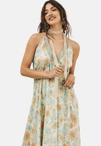 El vestido largo y ligero presenta un patrón floral en tonos pastel de turquesa, durazno y crema. Cuello halter con lazos texturizados y corte holgado.