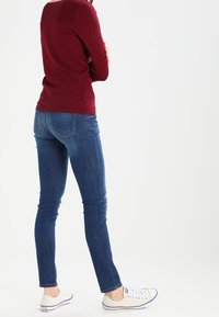Maglietta bordeaux a maniche lunghe con spalle testurizzate, abbinata a jeans skinny blu scuro e sneakers beige con suole bianche.