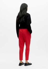 Rote, zulaufende Hose mit elastischem Bund, zwei Gesäßtaschen und glatter Textur, kombiniert mit einem schwarzen Langarmoberteil und schwarzen Schuhen.