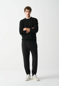 Schwarzer gestrickter Pullover mit gerippten Bündchen und Saum, kombiniert mit schwarzen Jogginghosen mit elastischen Bündchen; ergänzt durch beige Sneakers.
