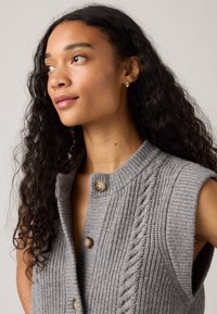 Gilet en tricot gris avec un col montant, présentant une fermeture à boutons et un motif en point de câble sur un côté. Côtes texturées sur l'ensemble.
