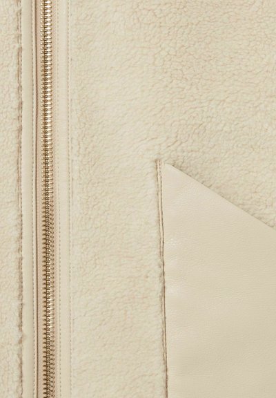 Il tessuto in pile beige chiaro presenta un aspetto tessurizzato, completato da un elegante accentuazione in pelle color crema e una cerniera in metallo dorato.