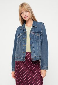 Jaqueta de ganga com botões à frente, dois bolsos no peito e uma cor azul desbotada, combinada com uma blusa verde-claro e uma saia roxa com bolinhas.