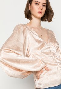 Blusa de cetim rosa pálido com padrão floral, decote em V profundo, mangas bufantes e botões frontais. Textura suave, ligeiramente ajustada na cintura.