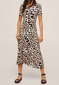 Femme portant une robe léopard à manches courtes et longueur midi, assortie à des sandales à brides noires, sur un fond uni.