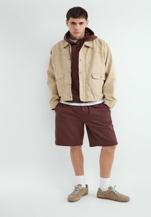 Jeune homme portant une veste beige par-dessus un sweat à capuche marron et un short, debout les mains dans les poches devant un fond blanc uni.